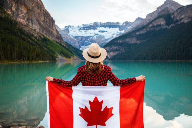 woman wearing red and black checkered dress shirt and beige fedora hat holding canada flag looking at lake